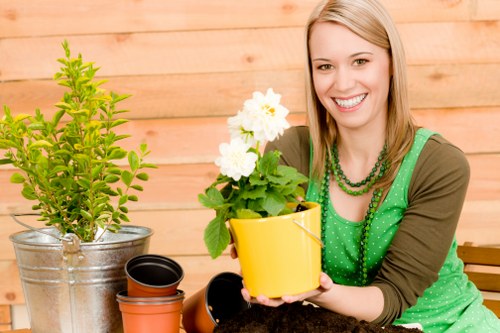 The image shows a young woman with light brown hair and a bright smile, sitting in an outdoor garden space with wooden paneling in the background. She is holding a yellow flower pot containing a small, healthy green plant with white flowers. On the table in front of her, there are various gardening pots made of different materials, including black and terracotta-colored pots, some of which are stacked or empty. The scene features a neatly maintained garden area with a lush, green lawn and some shrubbery, suggesting a well-kept outdoor space suitable for gardening and landscaping services in Lewisham. The natural lighting indicates a clear day, highlighting the vibrant colors of the plants and the woman's attire, creating a cheerful and inviting atmosphere linked to garden care and plant management activities consistent with local gardening services.