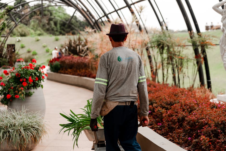 A gardener, dressed in a long-sleeved grey shirt with reflective stripes on the sleeves and wearing a dark hat, is walking through a well-maintained garden within a large greenhouse or conservatory. T