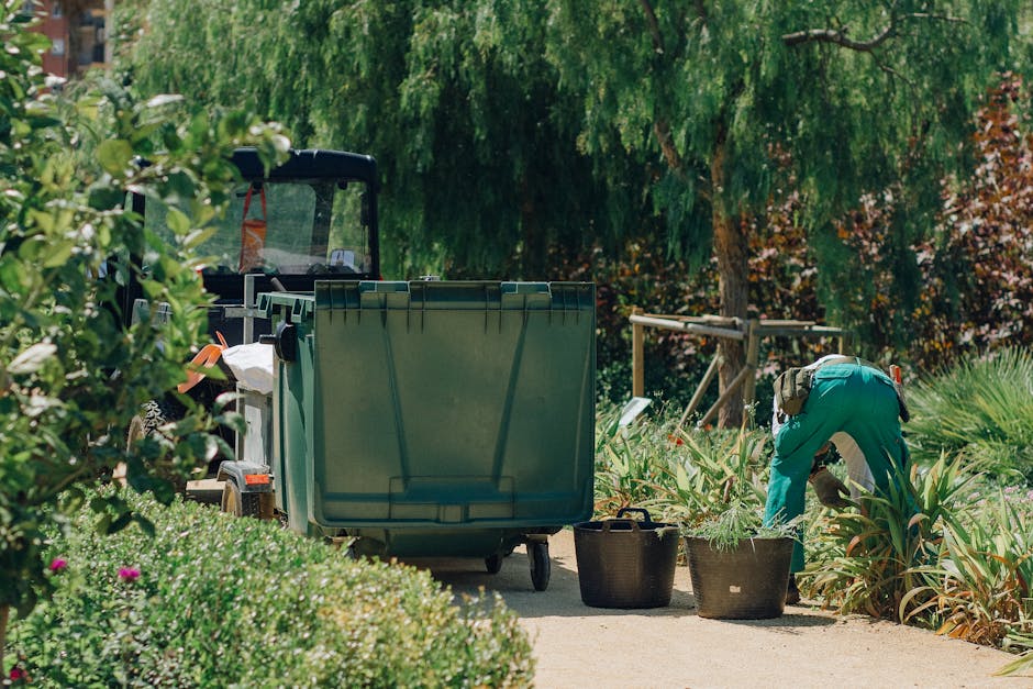 A gardener, dressed in a long-sleeved grey shirt with reflective stripes on the sleeves and wearing a dark hat, is walking through a well-maintained garden within a large greenhouse or conservatory. T