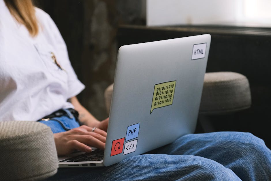 The image depicts a person seated outdoors on a beige cushioned chair, working on a silver laptop placed on their lap. The person is wearing a white shirt and blue jeans, with their left hand resting